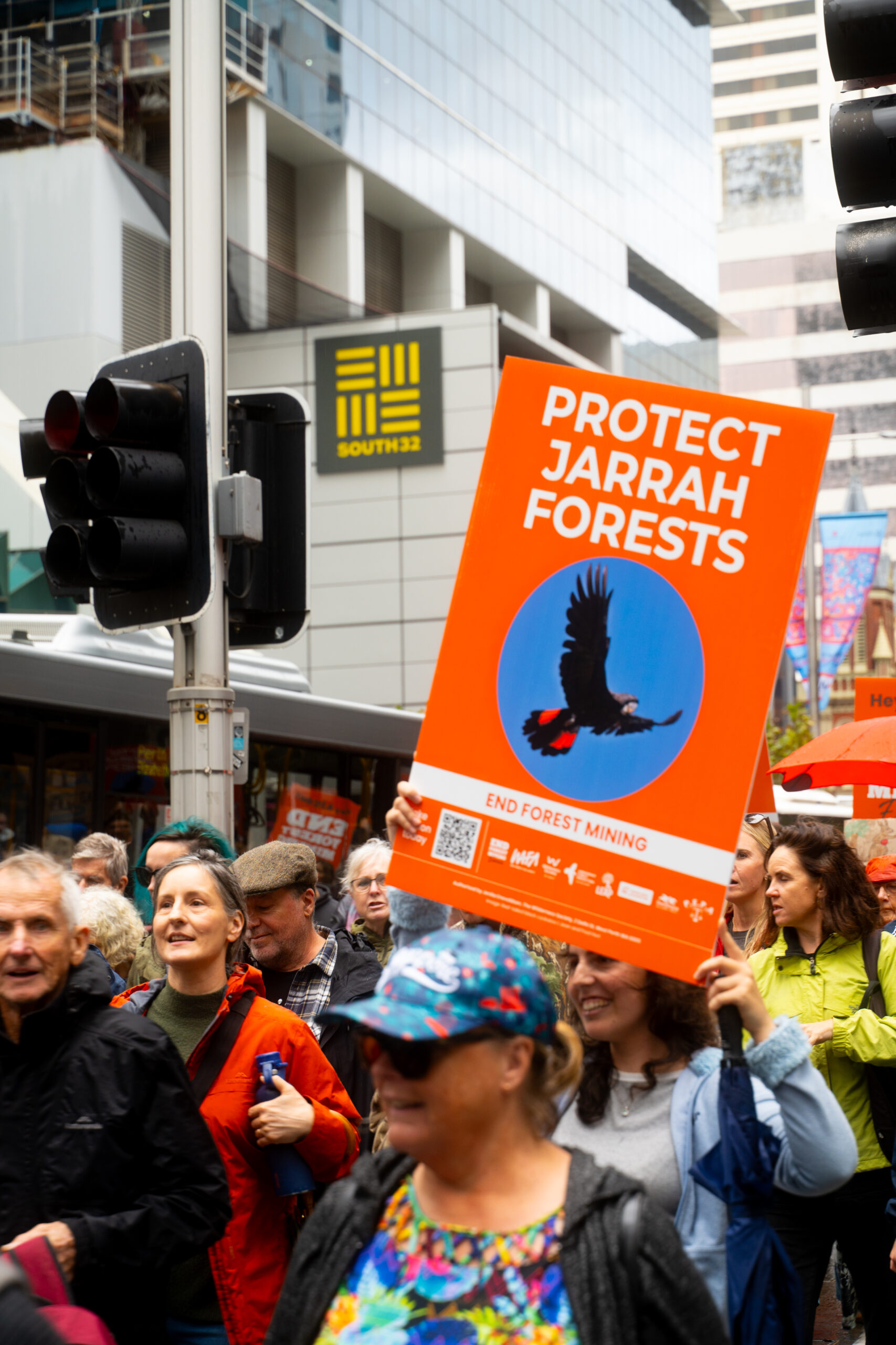A person in a crowd holding a sign reading "Protect Jarrah Forests" with an image of a black cockatoo.