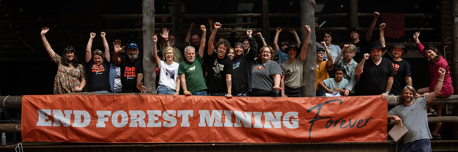 A group of people with their fists in the air behind an orange banner that says End Forest Mining Forever